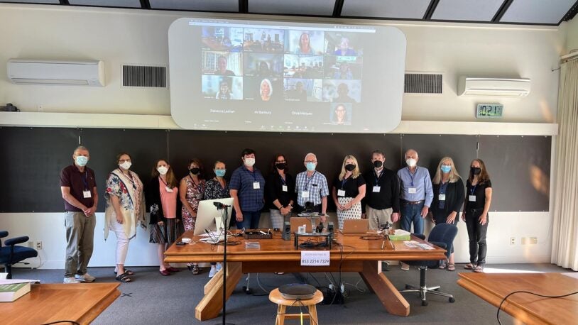 13 people standing in one row in the conference room. All are wearing masks and lanyards. Behind them, there are 14 people on the projector participating in the meeting virtually.
